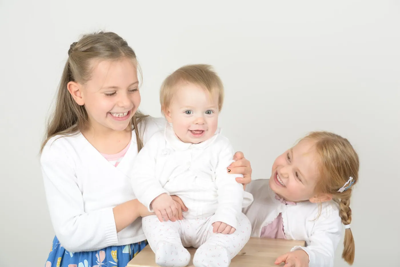 Deux jeunes sœurs riant avec leur petit frère bébé, photographiés sur fond clair au studio