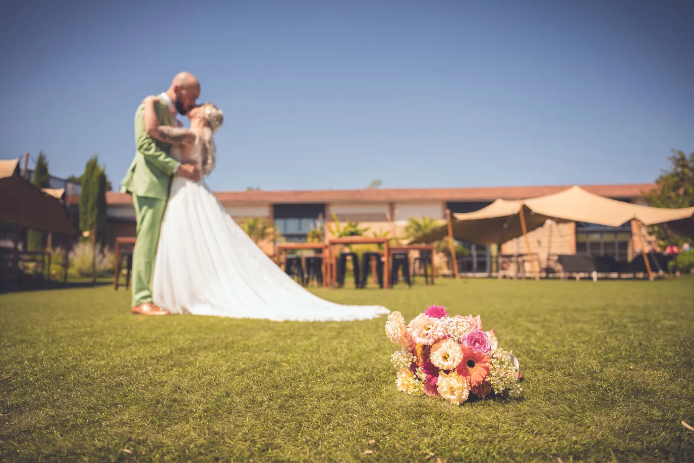 Les mariés s'embrassent dans le jardin de leur réception, le bouquet posé au premier plan sur la pelouse