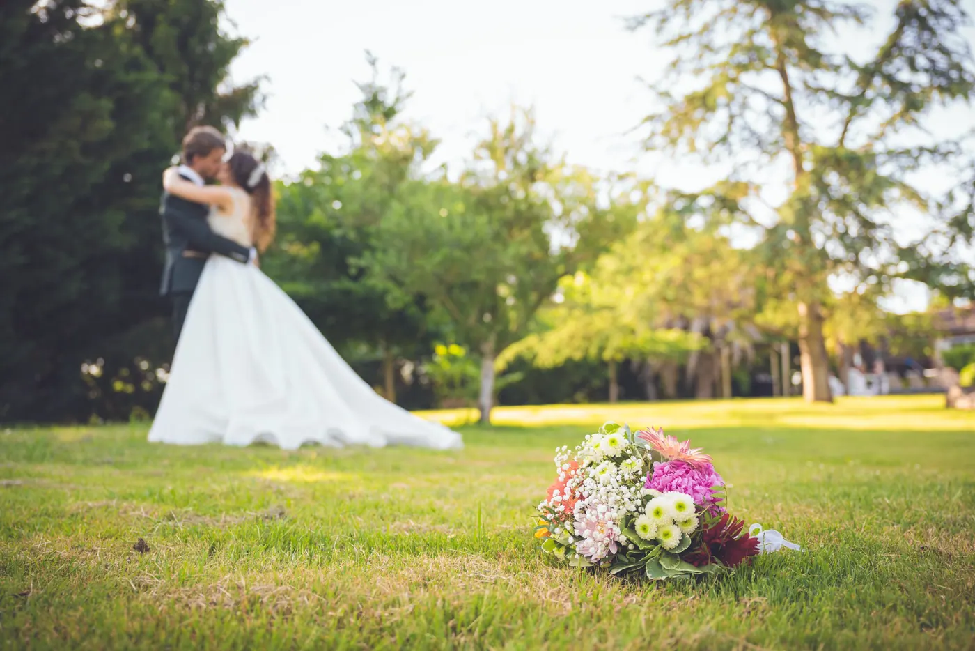Le bouquet posé dans l'herbe au premier plan, les mariés enlacés en arrière-plan dans le parc